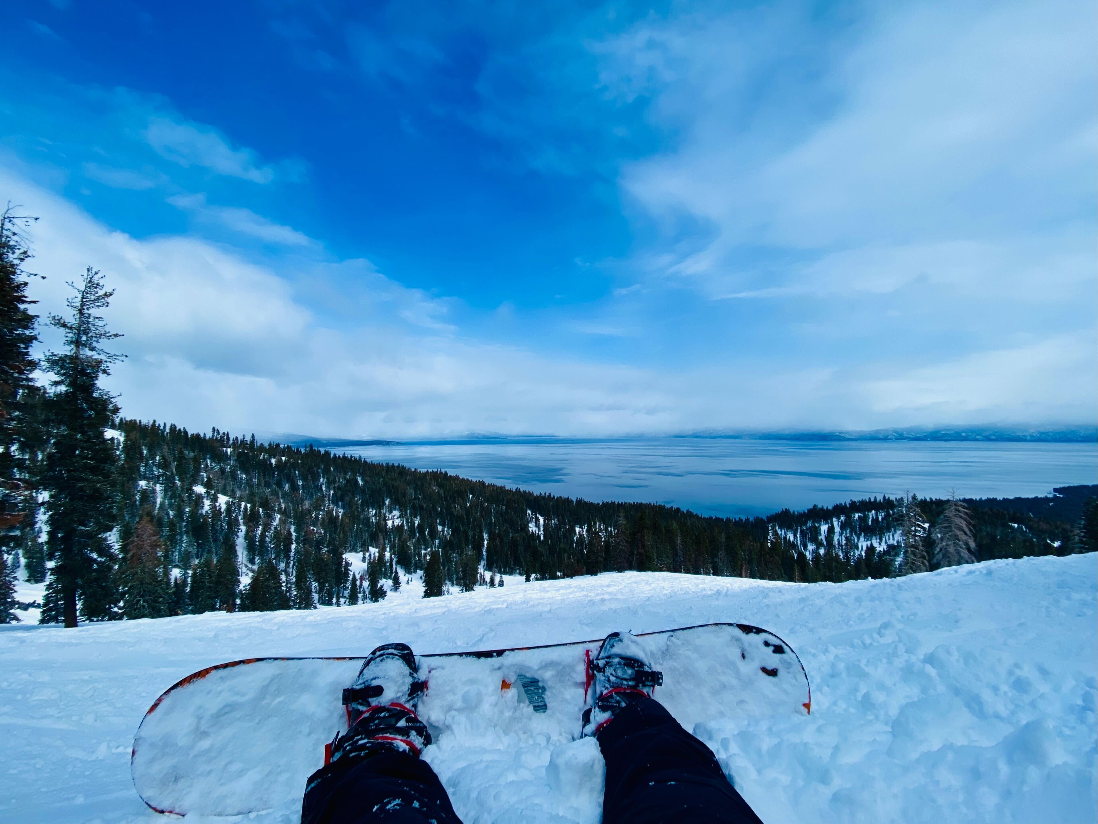 snowboard on top of a hill overlooking snow-covered hills, pine trees, and a clear blue lake in the distance 