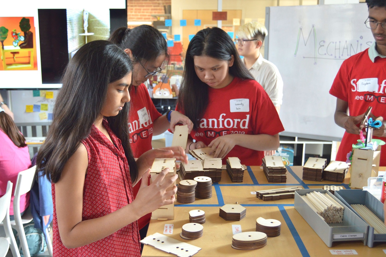 picture of a group of students looking at wooden supplies layed out on a table that they gathered to build their mechanisms 
