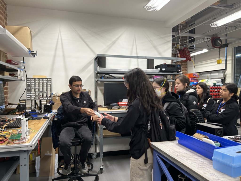 picture of a group of students gathering around Ahad, who is passing around a circuit board to show students his research